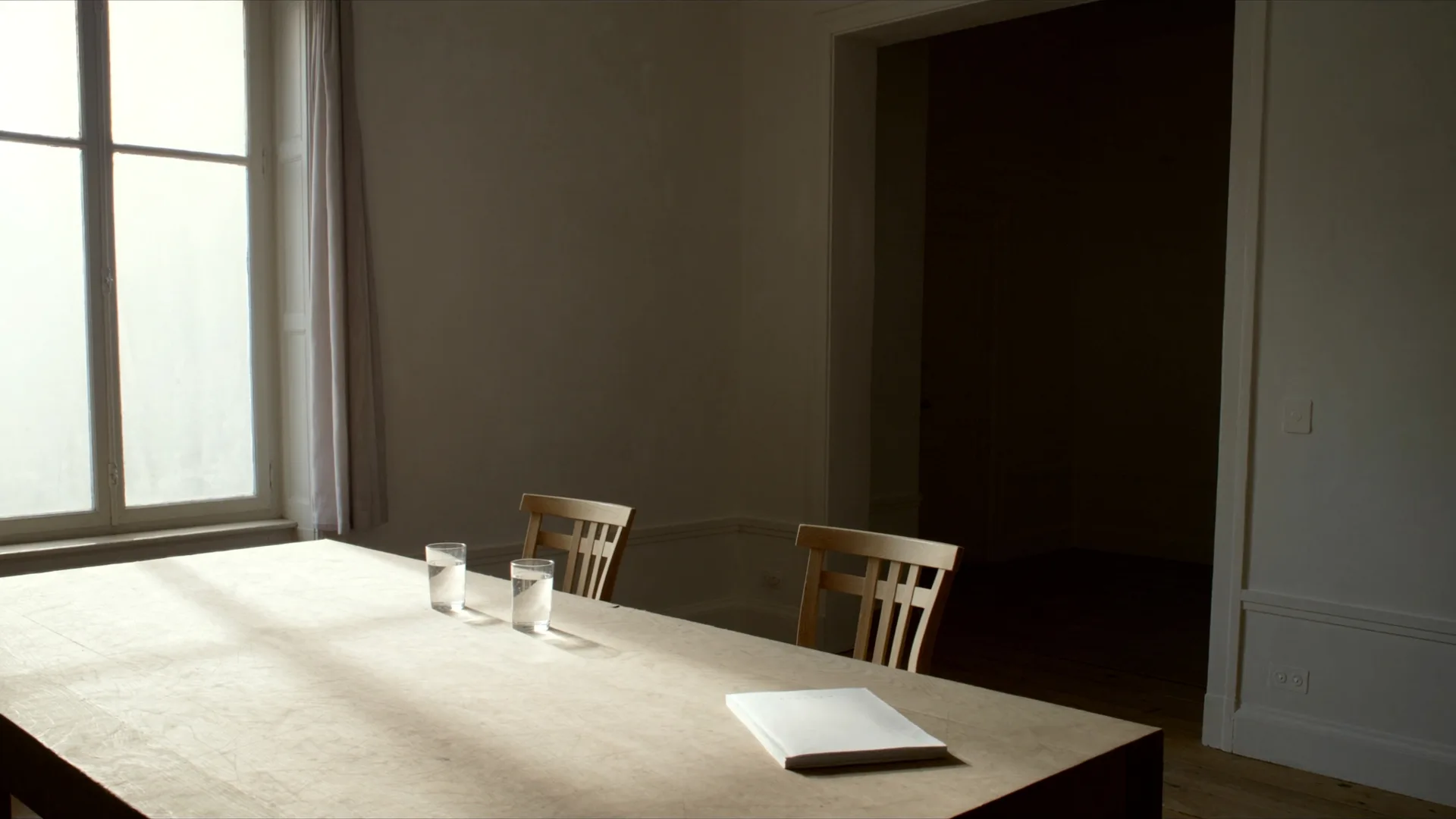 Twee lege stoelen aan een houten tafel, twee glazen water en een blanco schrift.
